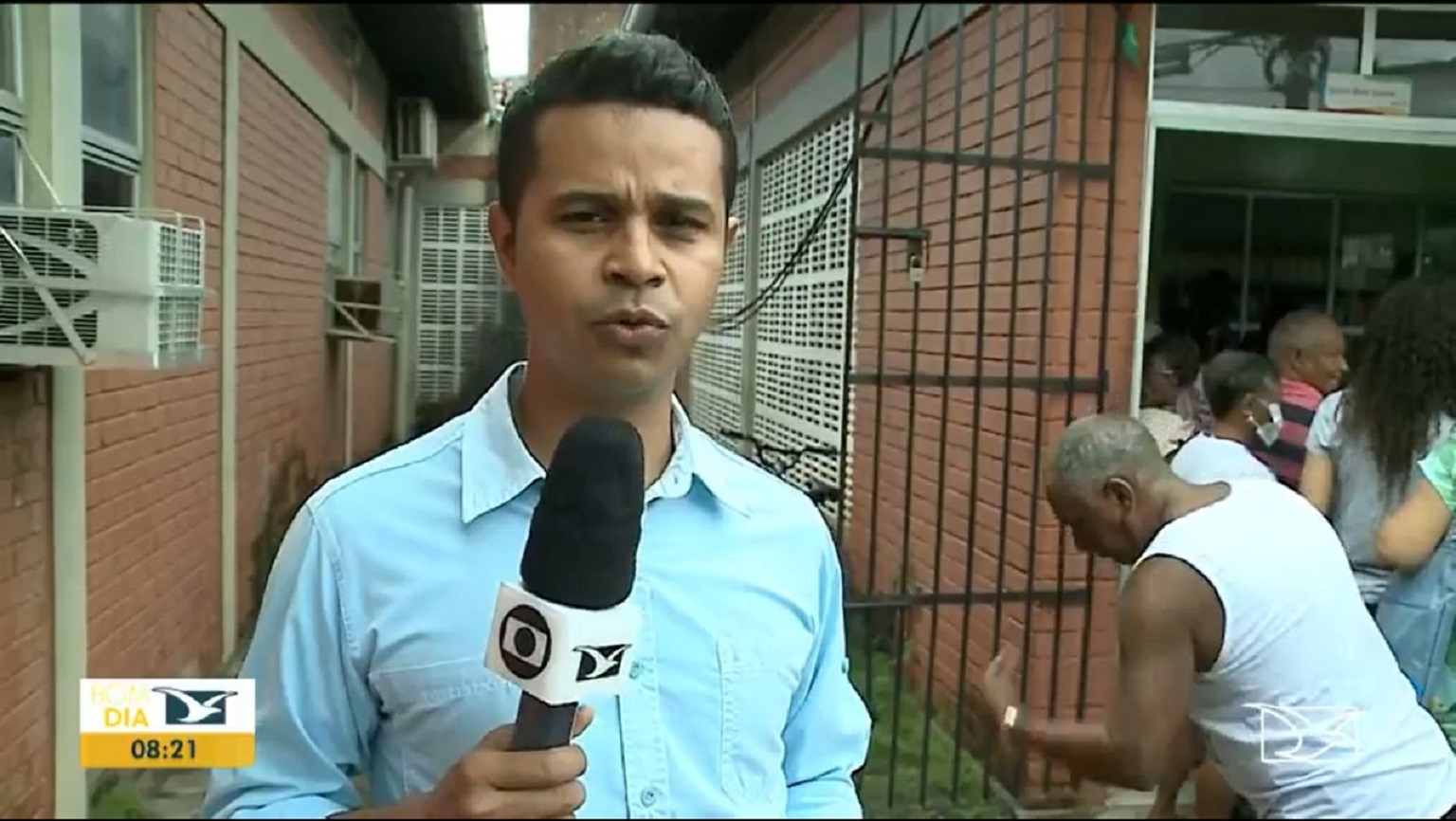 Repórter Douglas Pinto durante link do Bom Dia Mirante; ao fundo, idoso assoa o nariz em fila de vacinação
